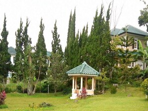 Um gazebo com telhado verde, rodeado por vegetação exuberante e árvores altas.