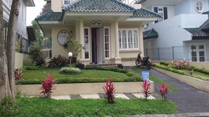 Une maison à deux étages, avec un toit recouvert de tuiles, un porche à l’avant et un jardin où poussent des plantes aux fleurs rouges.