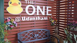 Un banc en bois avec une structure en métal, un mur sur lequel est affiché un panneau indiquant « THE ONE @ Udonthani », et des plantes en pot.