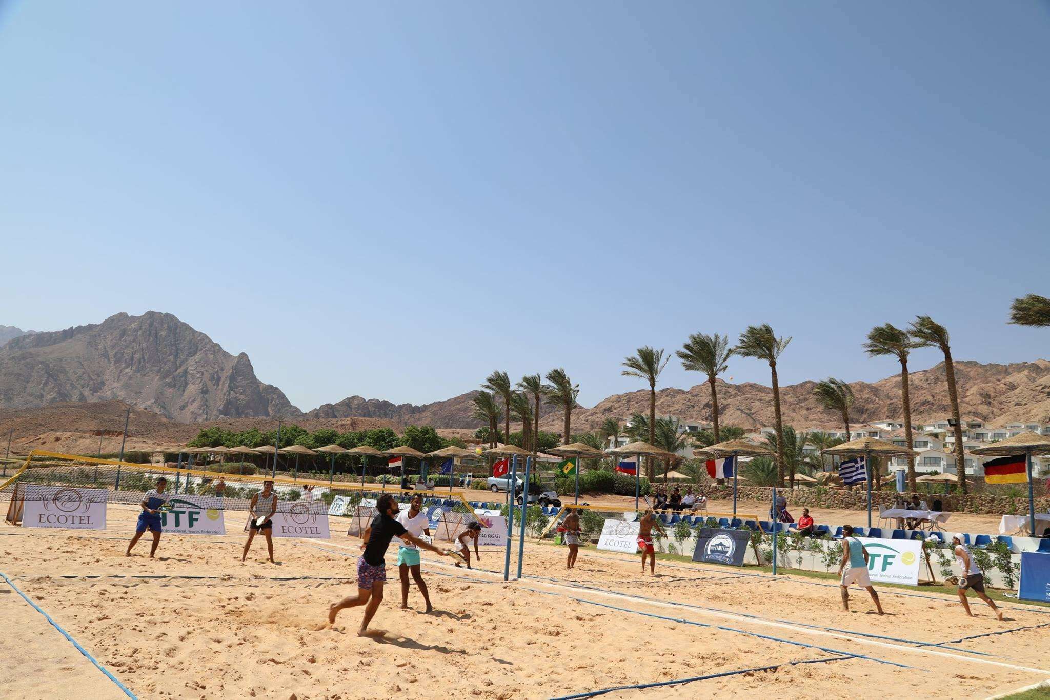 A beach volleyball court with players, palm trees, and mountains in the background.