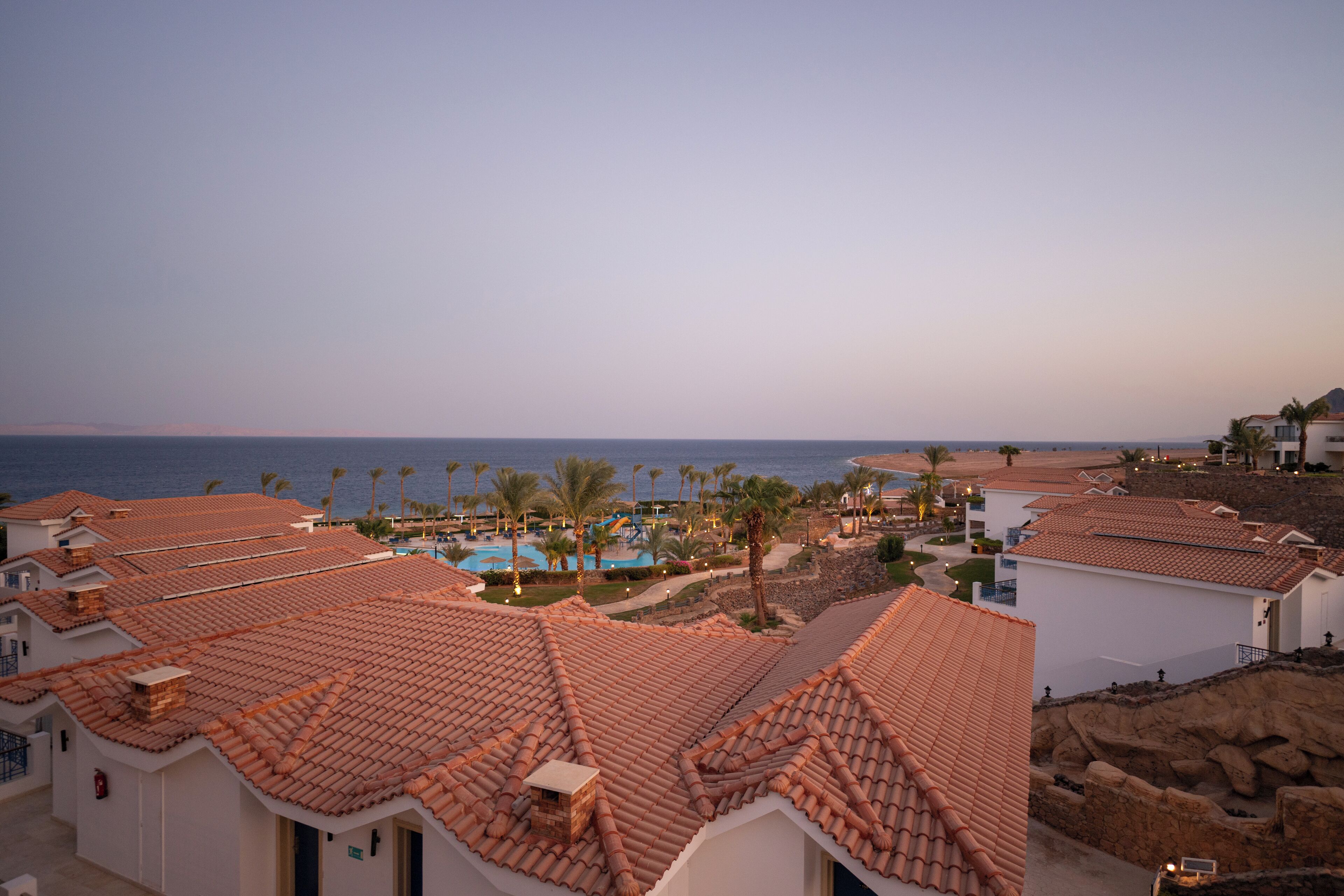A coastal resort with terracotta-tiled buildings, a swimming pool, and palm trees.