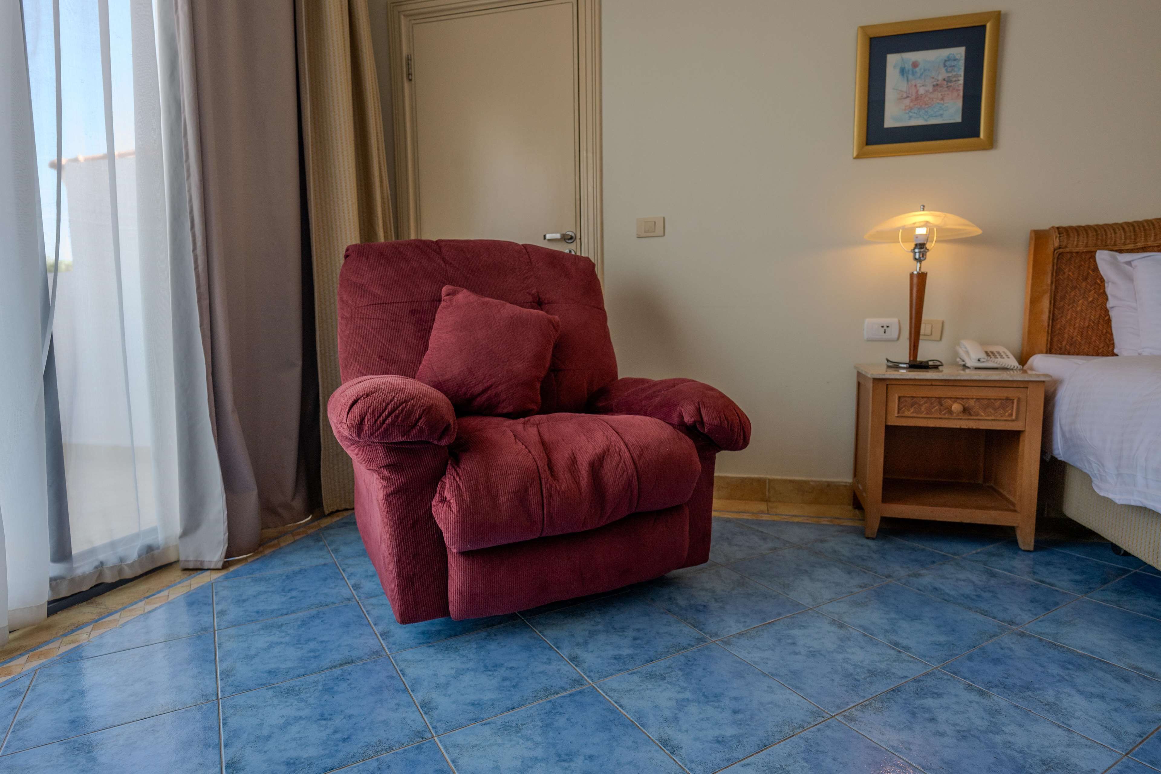A room with a red armchair, a wooden nightstand with a lamp, a bed, and a door.