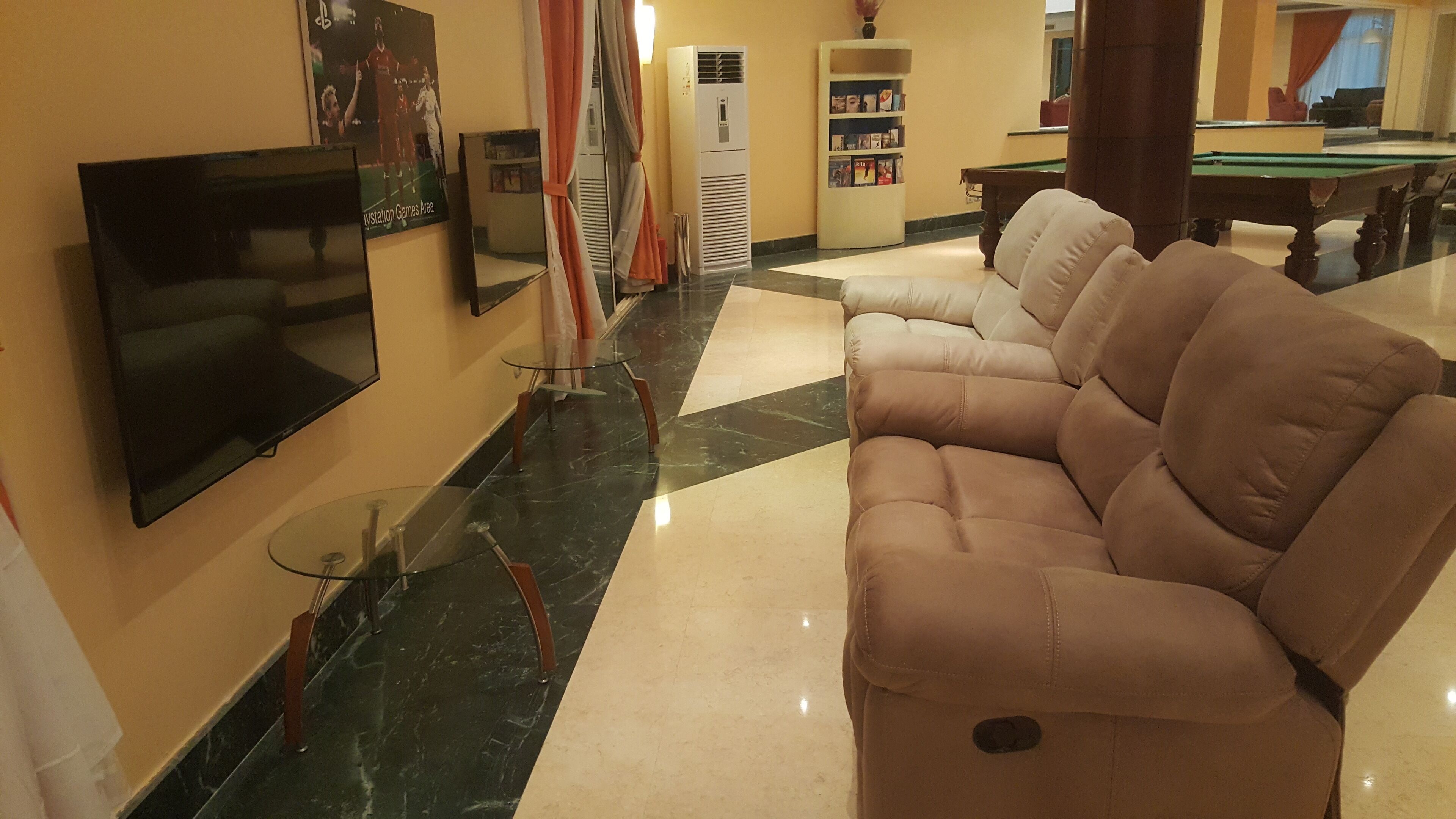 A living room with a flat-screen TV, a beige sectional sofa, a glass coffee table, a pool table, and a bookshelf.