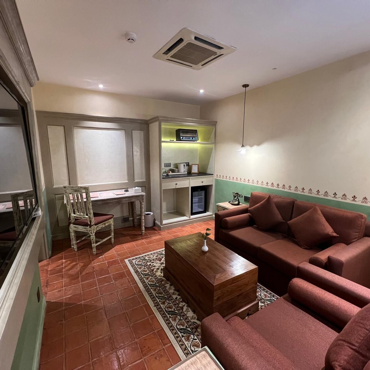 A living room with a sofa, a coffee table, a chair, a small kitchen area with a refrigerator, and a decorative wall border.