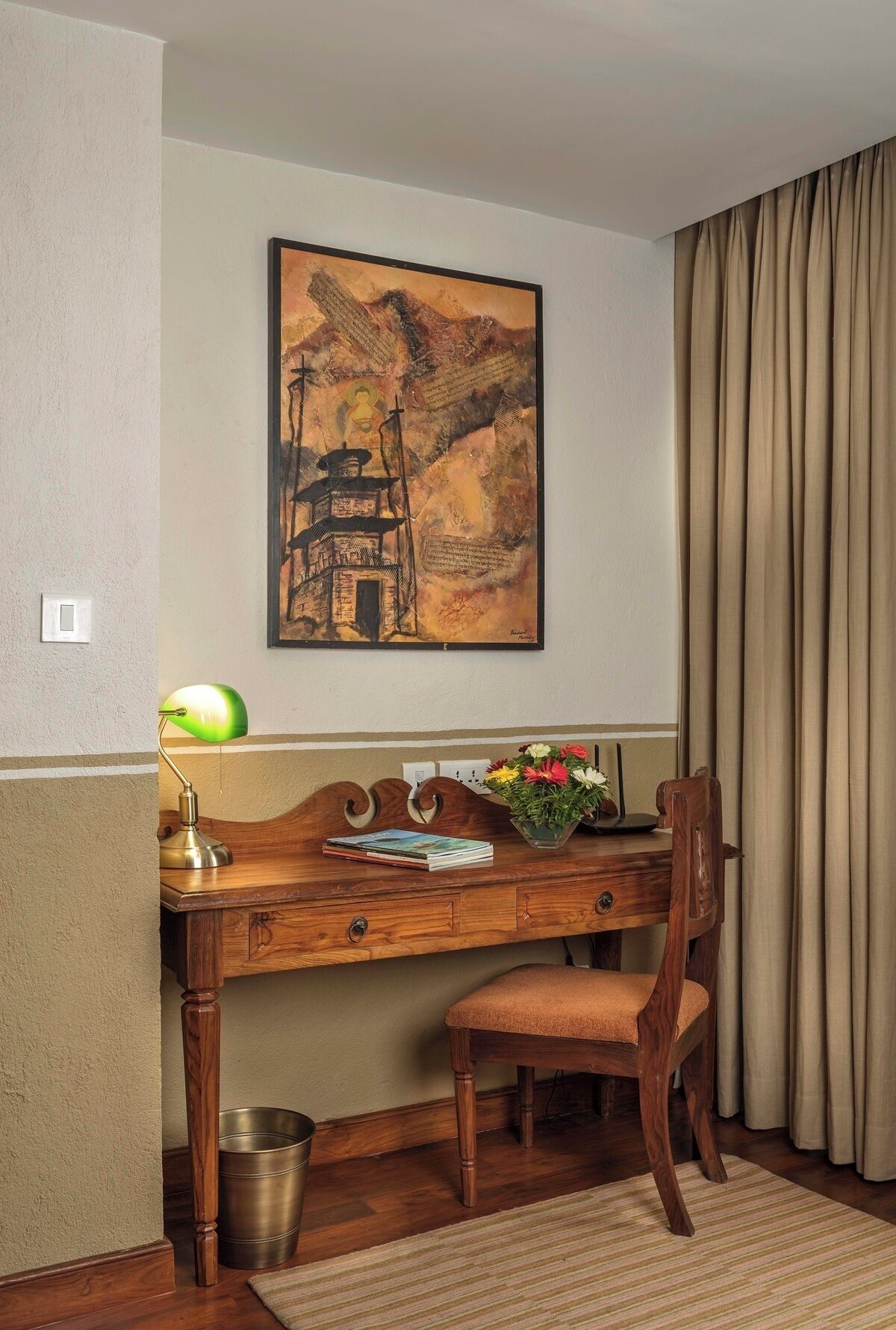 Habitación de hotel con escritorio de madera, una silla, una lámpara, un cuadro enmarcado, un jarrón con flores y una cortina.