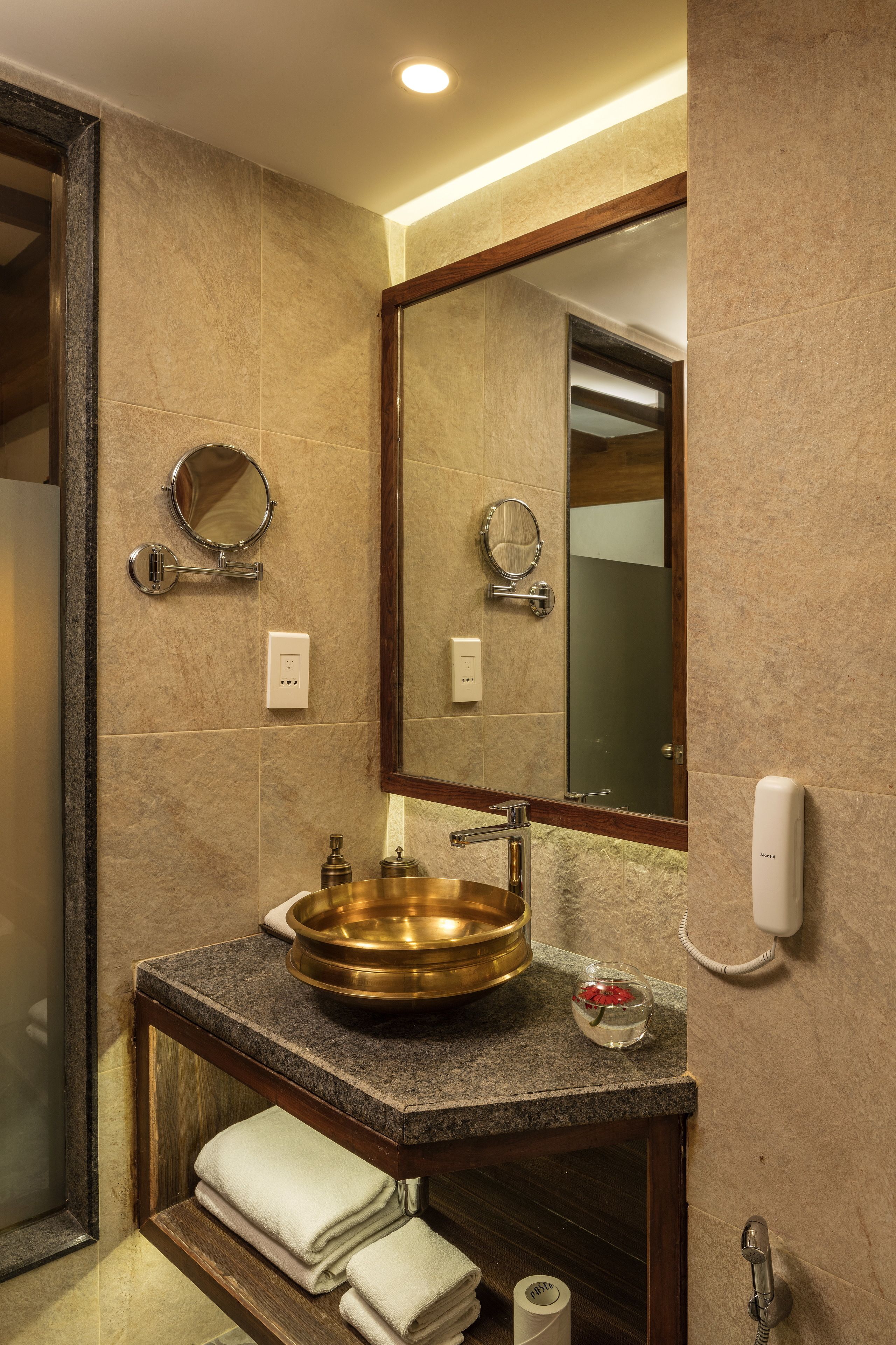 A bathroom with a granite countertop, a brass sink, a round mirror, a towel rack, and a hand dryer.