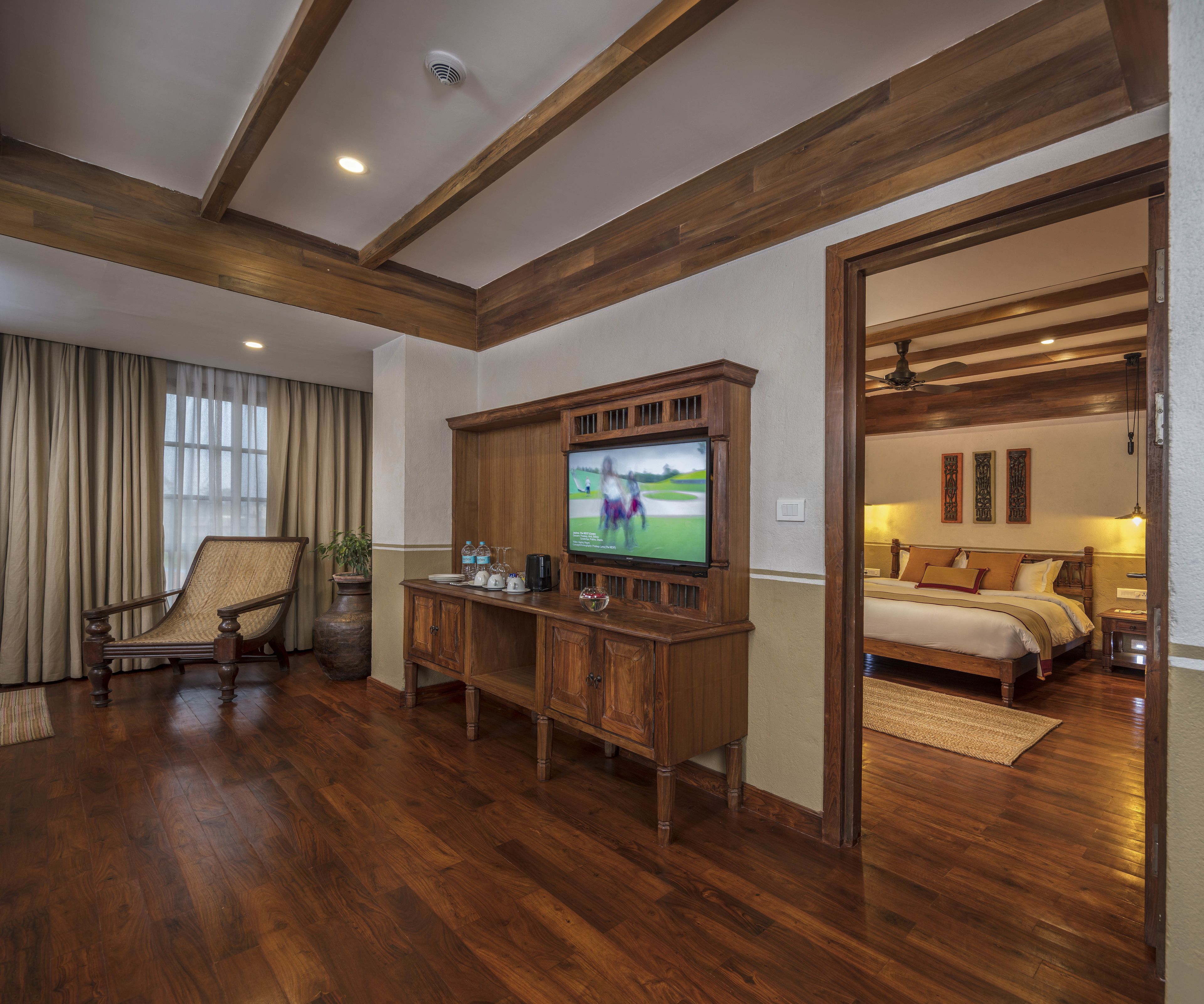 Ein Zimmer mit einem Holz-Entertainment-Center, einem Fernseher, einem großen Fenster mit Vorhängen, einem Holzstuhl, einem Bett mit Kissen und einem Deckenventilator.