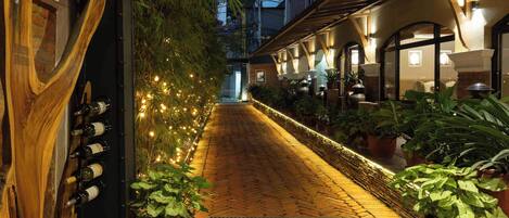 A narrow, brick-paved pathway lined with potted plants and string lights, leading to a building with arched windows and outdoor seating.