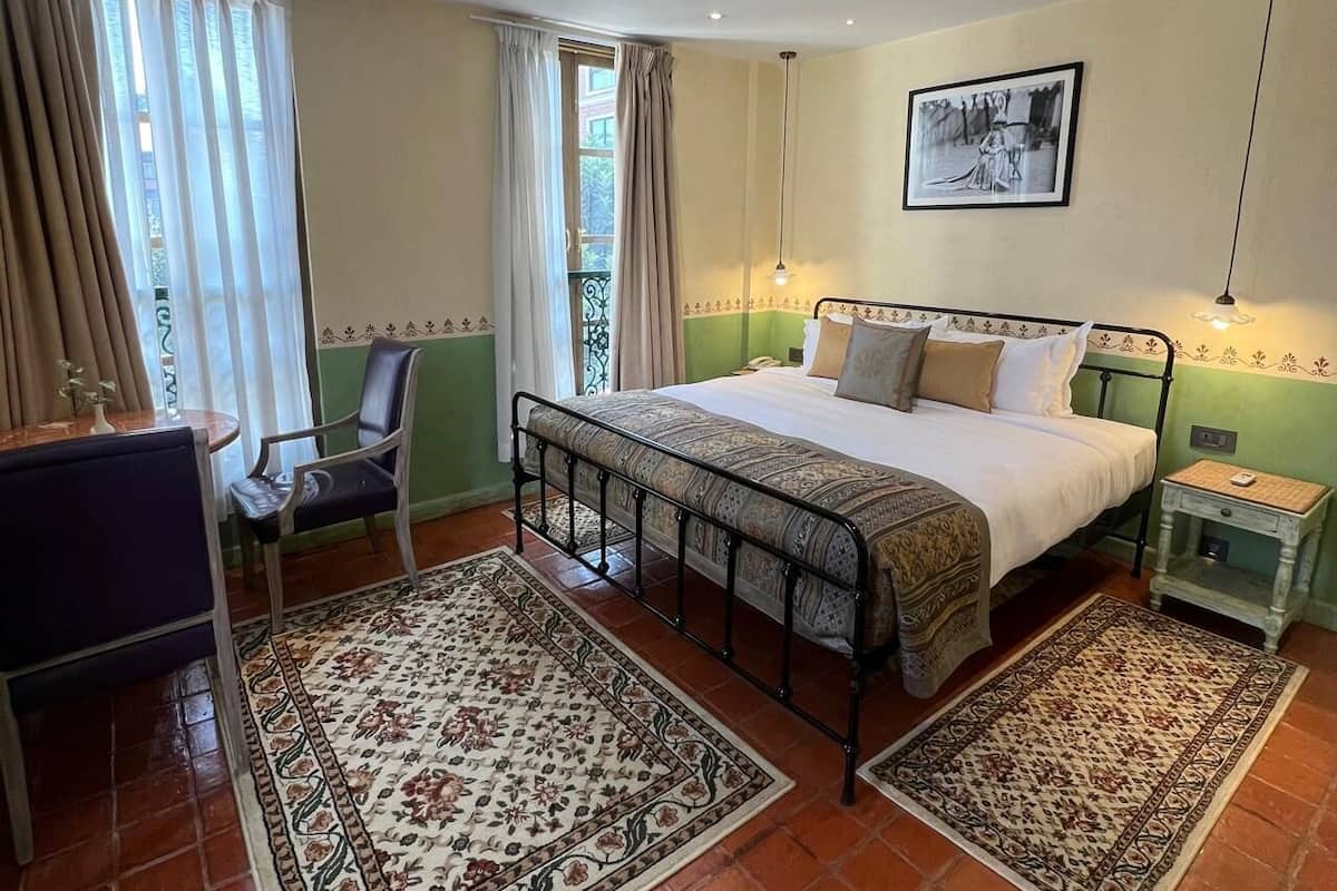 Un dormitorio con una cama, una silla, una mesita de noche, un cuadro en la pared y una ventana con cortinas.