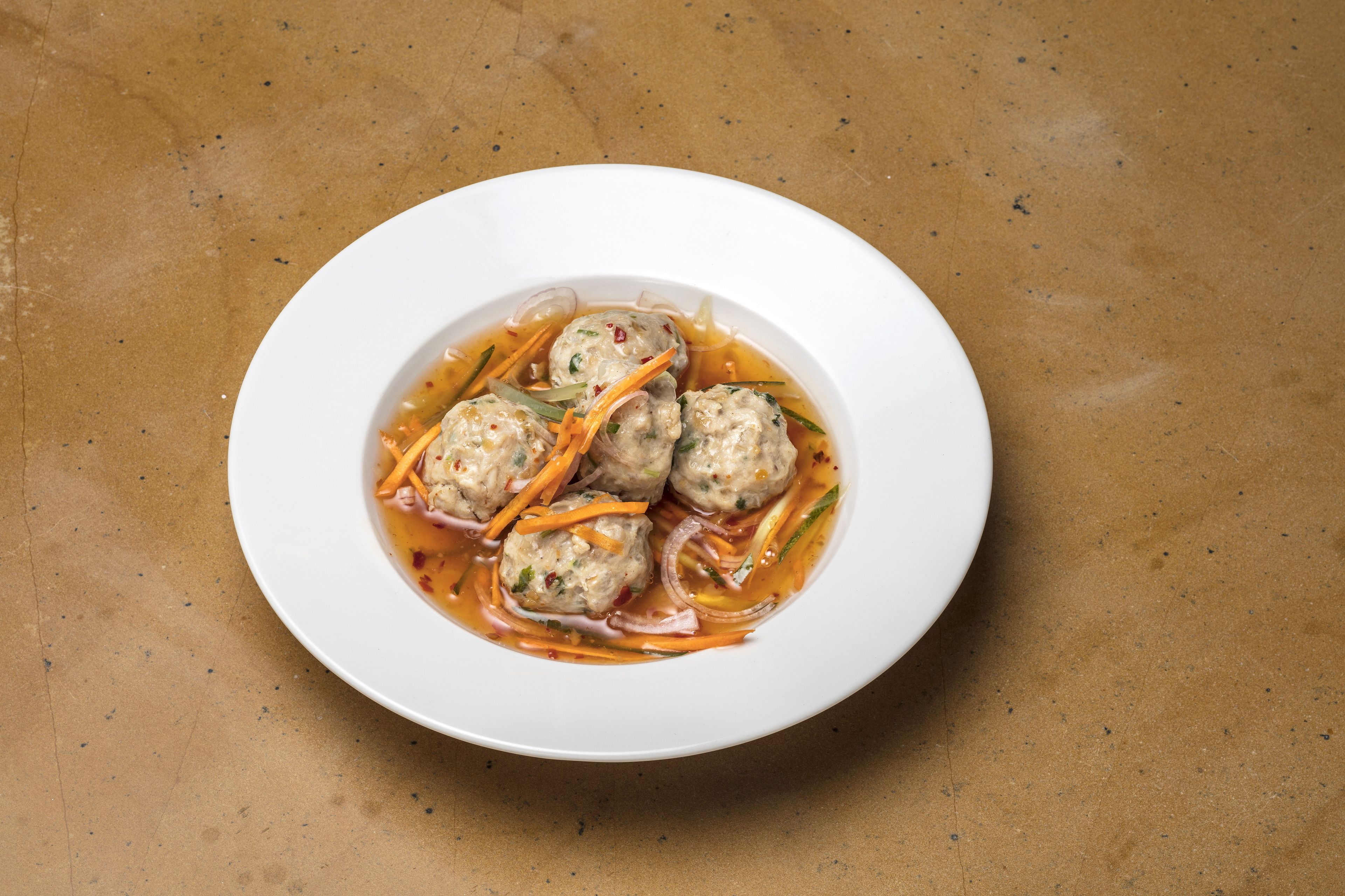 A white plate with four meatballs in a sauce, garnished with carrot strips and green herbs, on a textured brown surface.