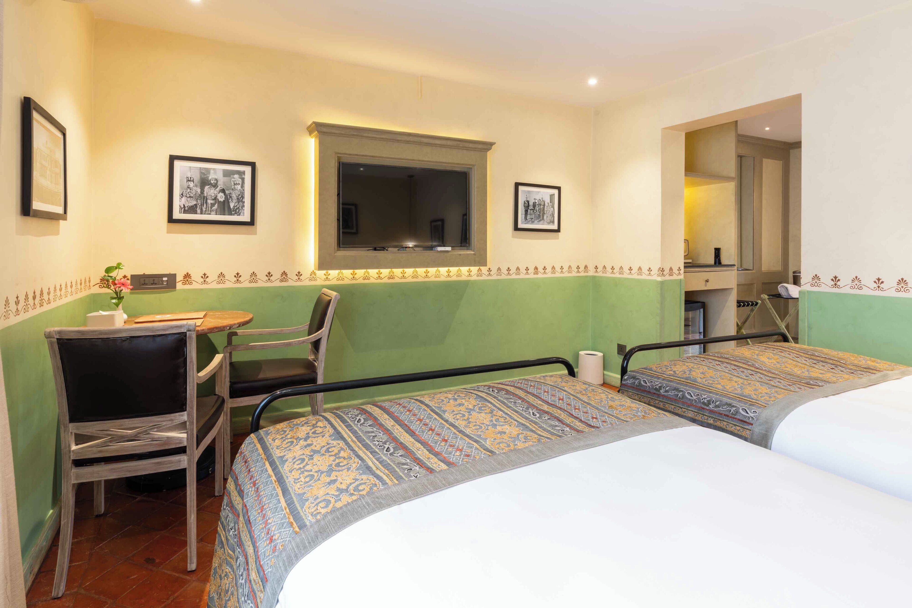 A hotel room with two beds, a small table, a chair, a window, and framed pictures on the walls.