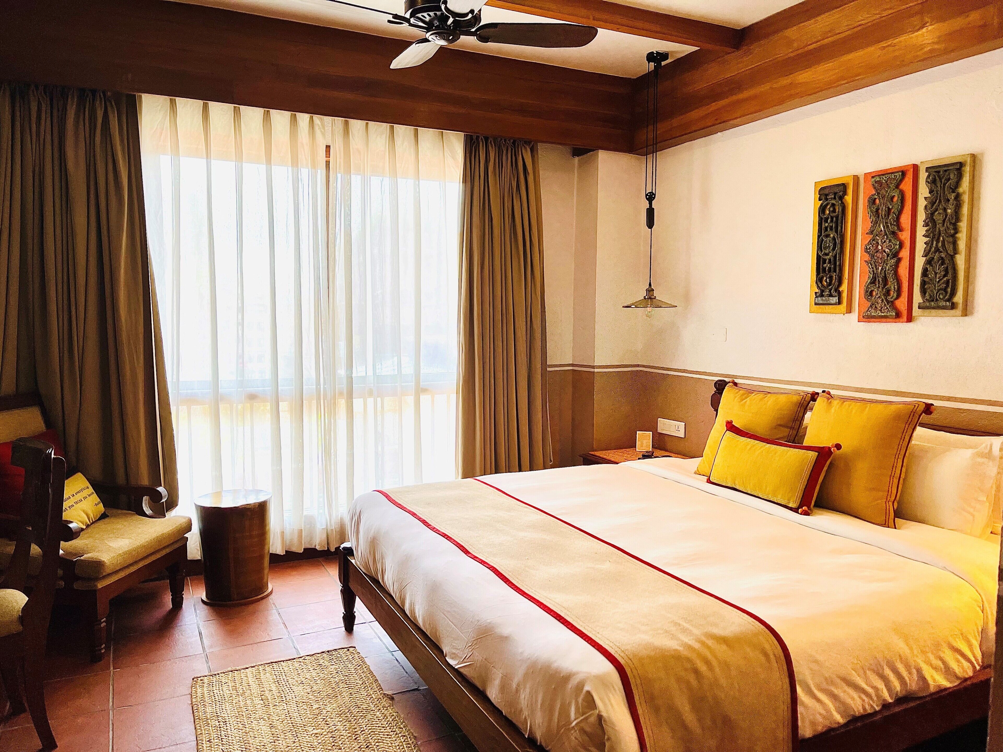 A hotel room with a large bed, wooden headboard, a ceiling fan, a bedside table with a lamp, a chair, and a view of the outdoors through sheer curtains.