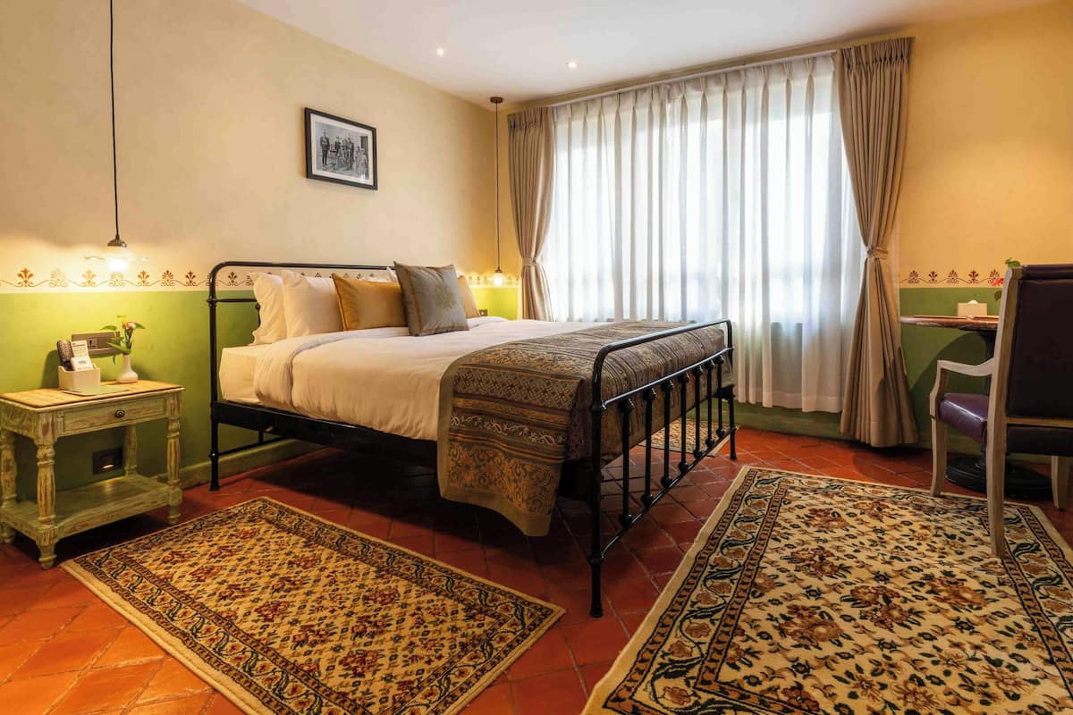 Un dormitorio con una cama, dos mesitas de noche, una silla, un escritorio y un ventanal con cortinas.