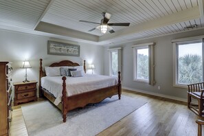 A bedroom with a wooden four-poster bed, two bedside lamps, a ceiling fan, and large windows with a view of trees. - Cassina Palms (Hilton Head Island)