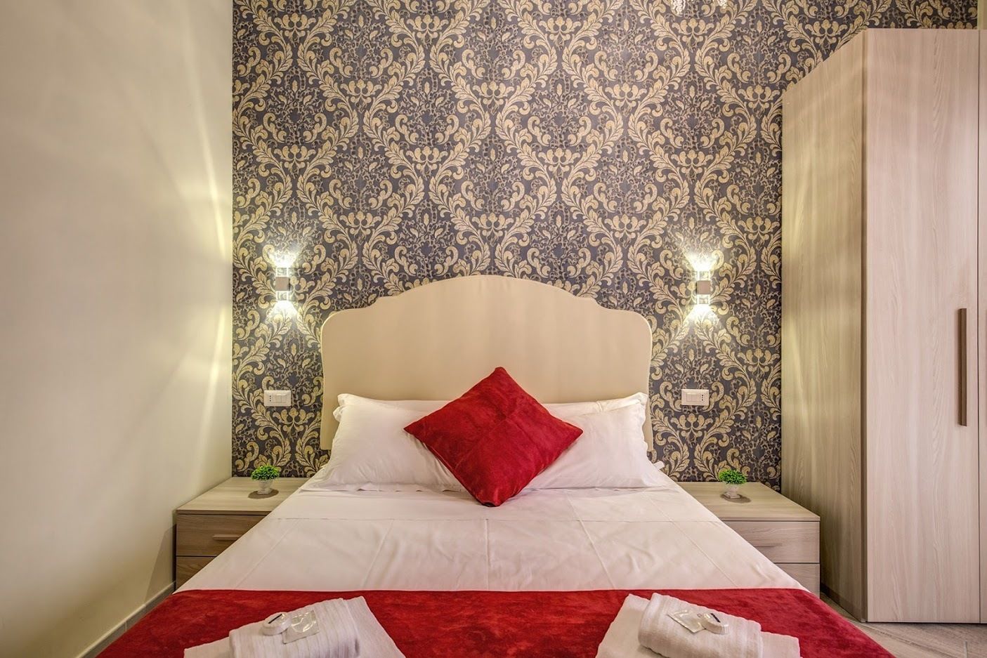 A neatly made bed with white linens and a red pillow, two bedside lamps, a wooden wardrobe, and patterned wallpaper.
