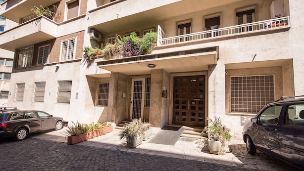 Un immeuble à plusieurs étages doté d’une entrée centrale, de balcons ornés de plantes et d’une voiture garée devant.