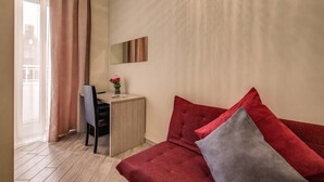 Une pièce comprenant un bureau en bois, une chaise, un canapé rouge avec des coussins gris, une petite table sur laquelle est posé un vase de fleurs, et une fenêtre avec des rideaux.