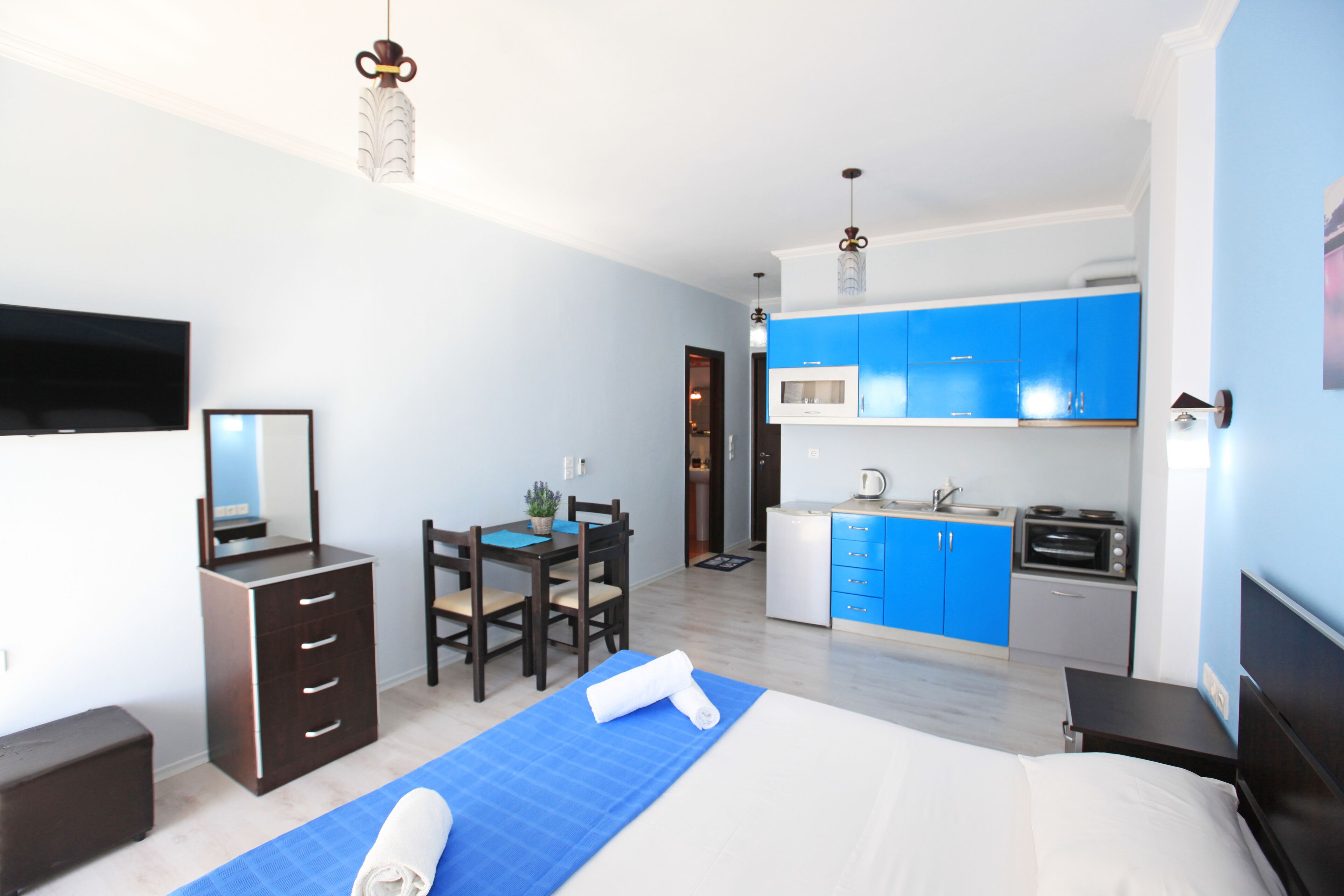 Quarto de hotel moderno com cama, escrivaninha, cadeira, cômoda e cozinha compacta.