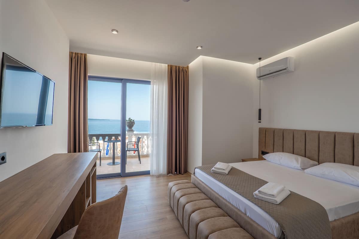 A modern hotel room with a large bed, a desk, and a TV. There is a balcony with a view of the sea.