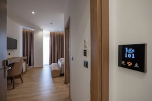 Quarto de hotel com piso de madeira, uma escrivaninha com cadeira, uma cama e uma televisão fixada na parede.