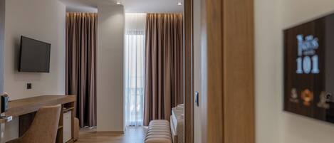 A modern hotel room with a wooden desk, a flat-screen TV, a sofa, and a large window with curtains.