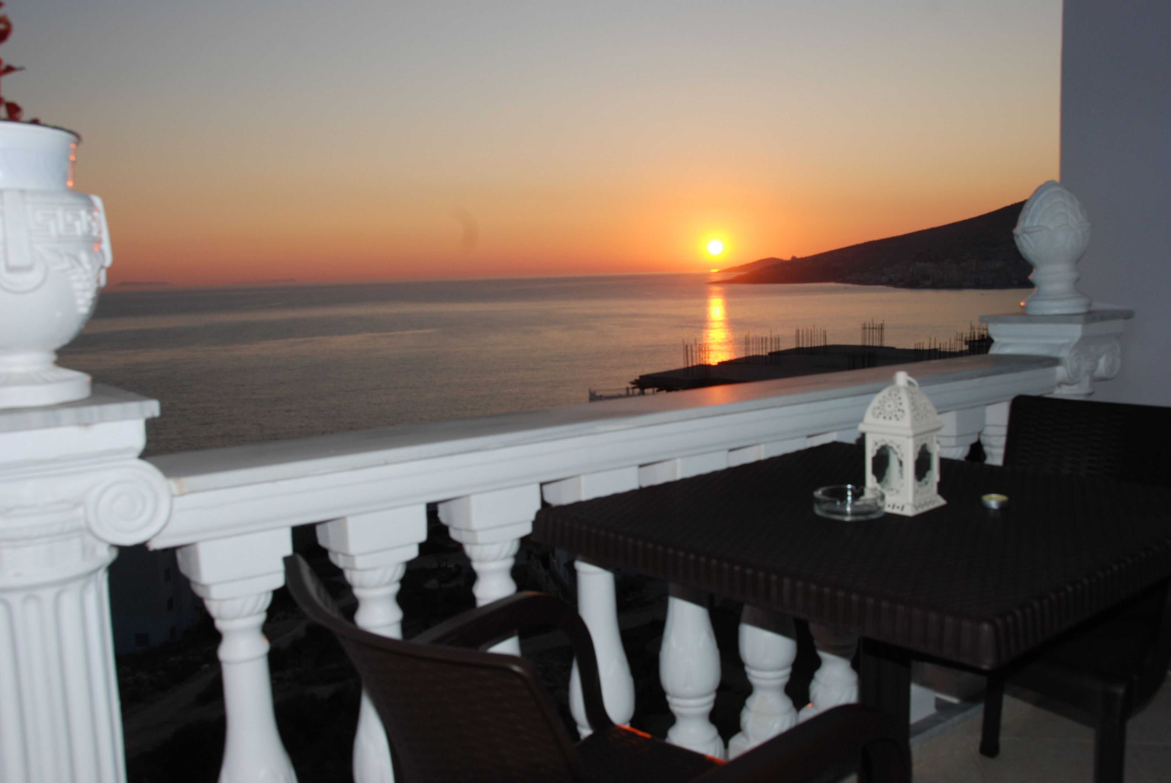 A balcony with a view of the sunset over the sea, a white balustrade, and a table with chairs.