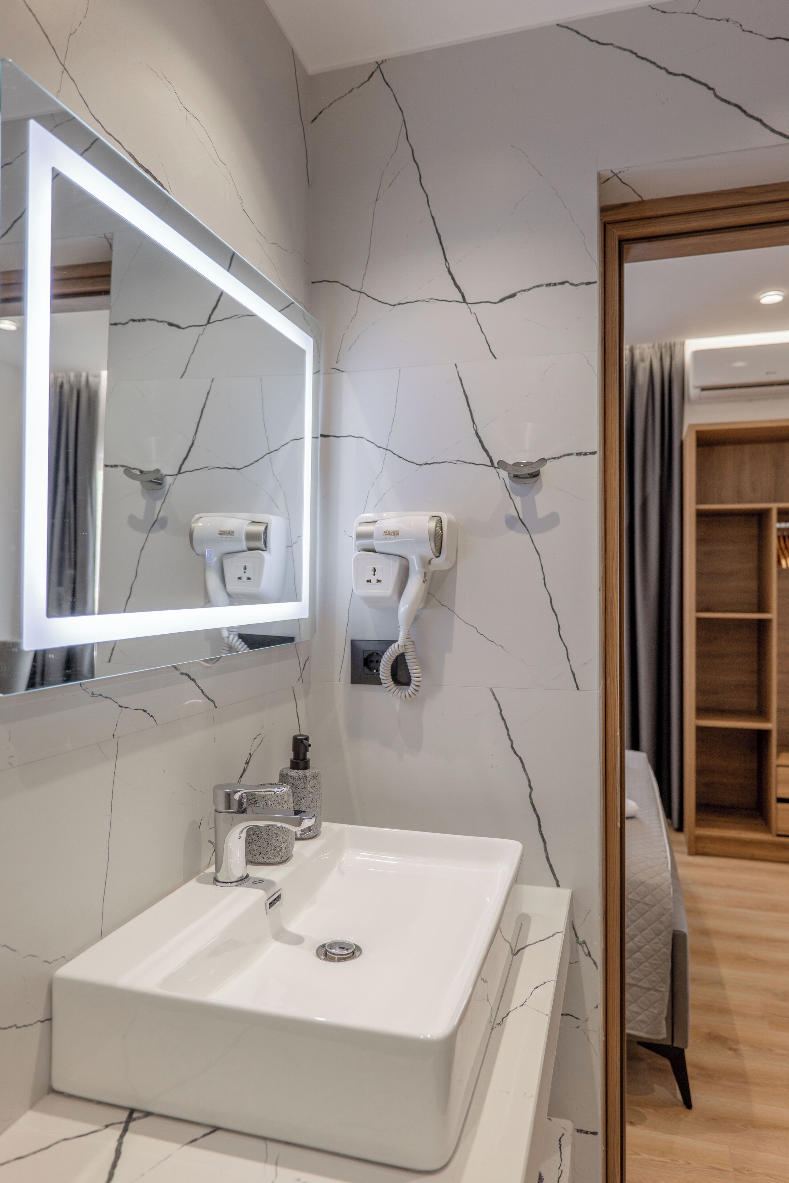 A modern bathroom with a white sink, a mirror with built-in lighting, and hair dryers mounted on the wall.