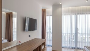 A modern hotel room with a wooden desk, a flat-screen TV, a sofa, and large windows with curtains. - Hotel Keos (Sarandë)