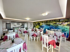 A dining area with tables set for guests, a kitchen in the background, and a mural of a river scene on the wall. - Hotel Keos (Sarandë)