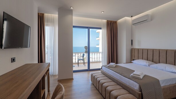 A modern hotel room with a large bed, a flat-screen TV, a wooden desk, and a balcony with a view of the sea. - Hotel Keos (Sarandë)