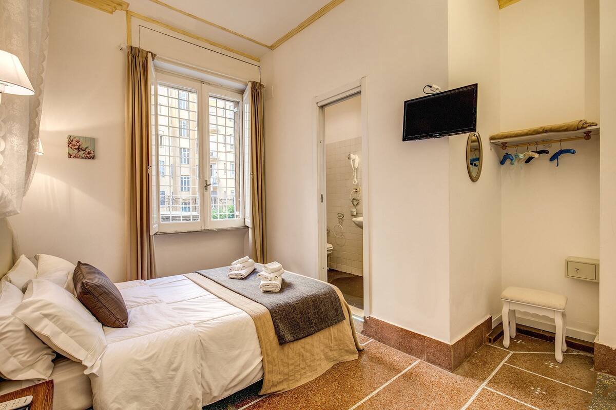 Habitación de hotel con cama, mesita de noche, silla, televisor y baño a la vista a través de una puerta abierta.