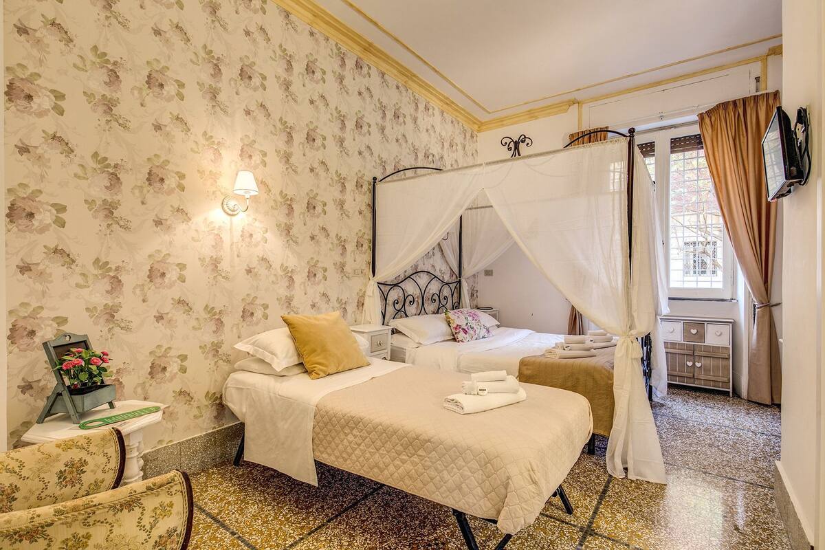 Un dormitorio con dos camas, papel tapiz floral, una mesita de noche y una ventana con cortinas.
