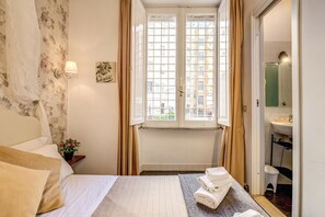 A hotel room with a bed, a window with curtains, a bedside table with a lamp, and a bathroom visible through an open door. - Bbdelux - Colosseo12 (Rome)