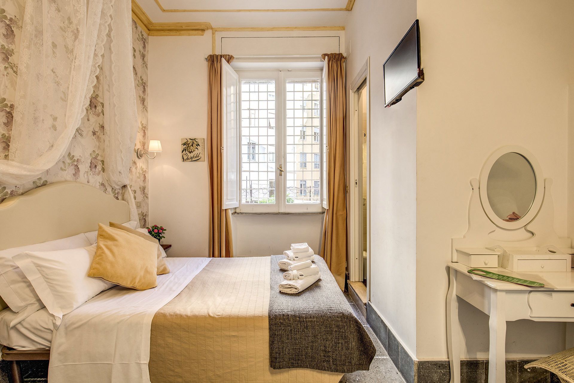 A hotel room with a bed, a desk with a mirror, a window with curtains, and a TV mounted on the wall.