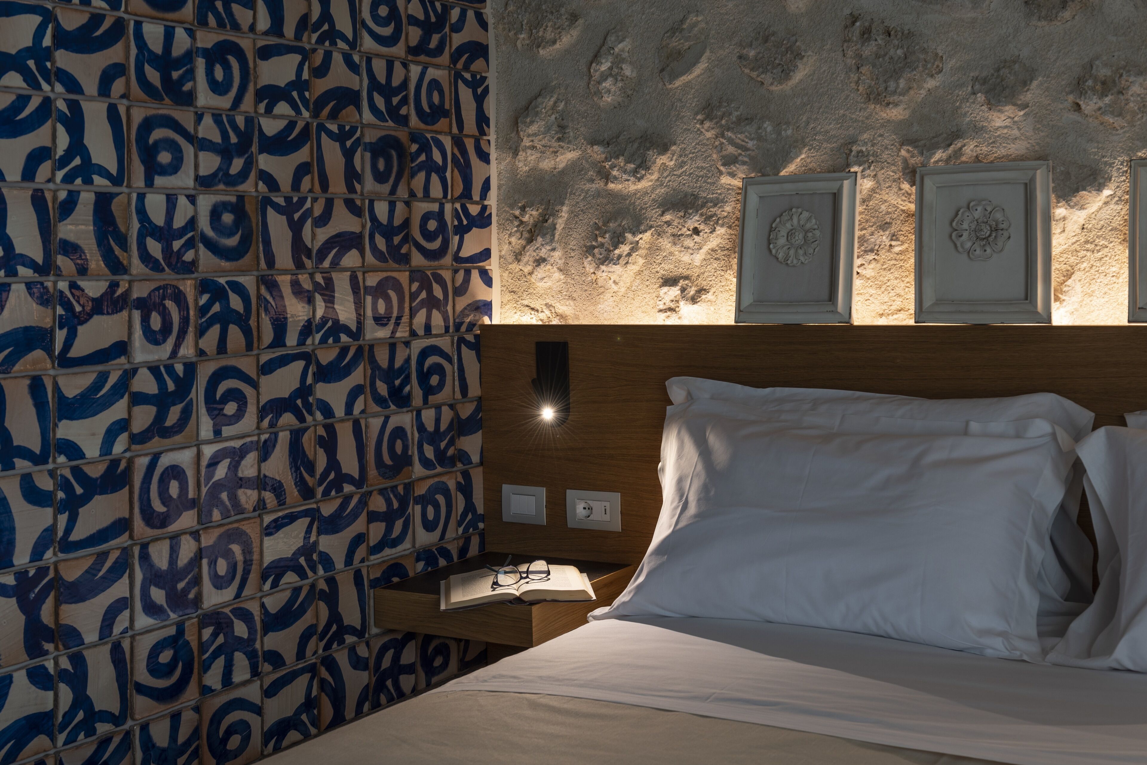 A hotel room with a patterned wall, a wooden headboard, a bedside table with a book and glasses, and two wall-mounted decorative items.