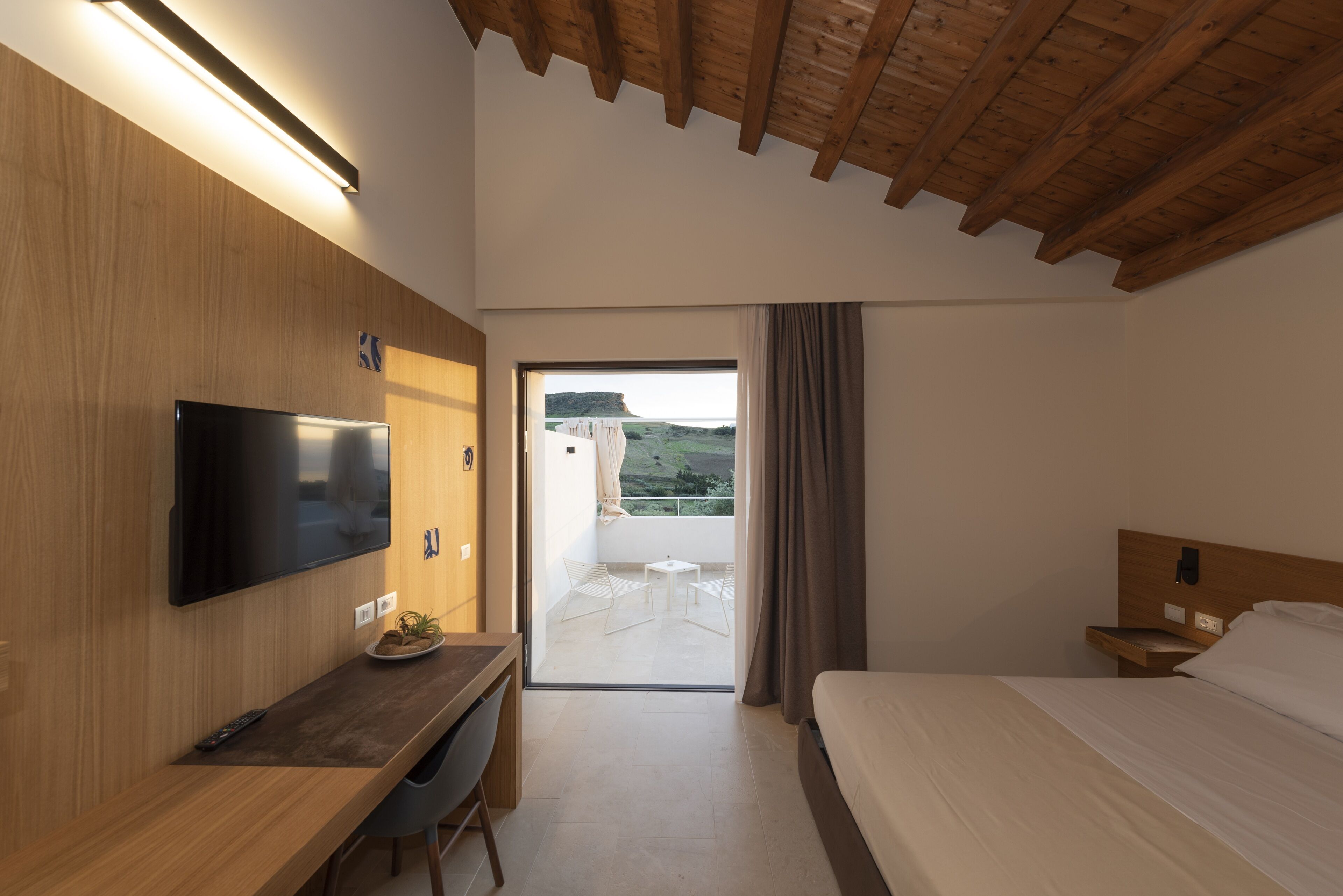 Een moderne hotelkamer met een flatscreen televisie, een houten bureau, een bed en een balkon met uitzicht op het landschap.