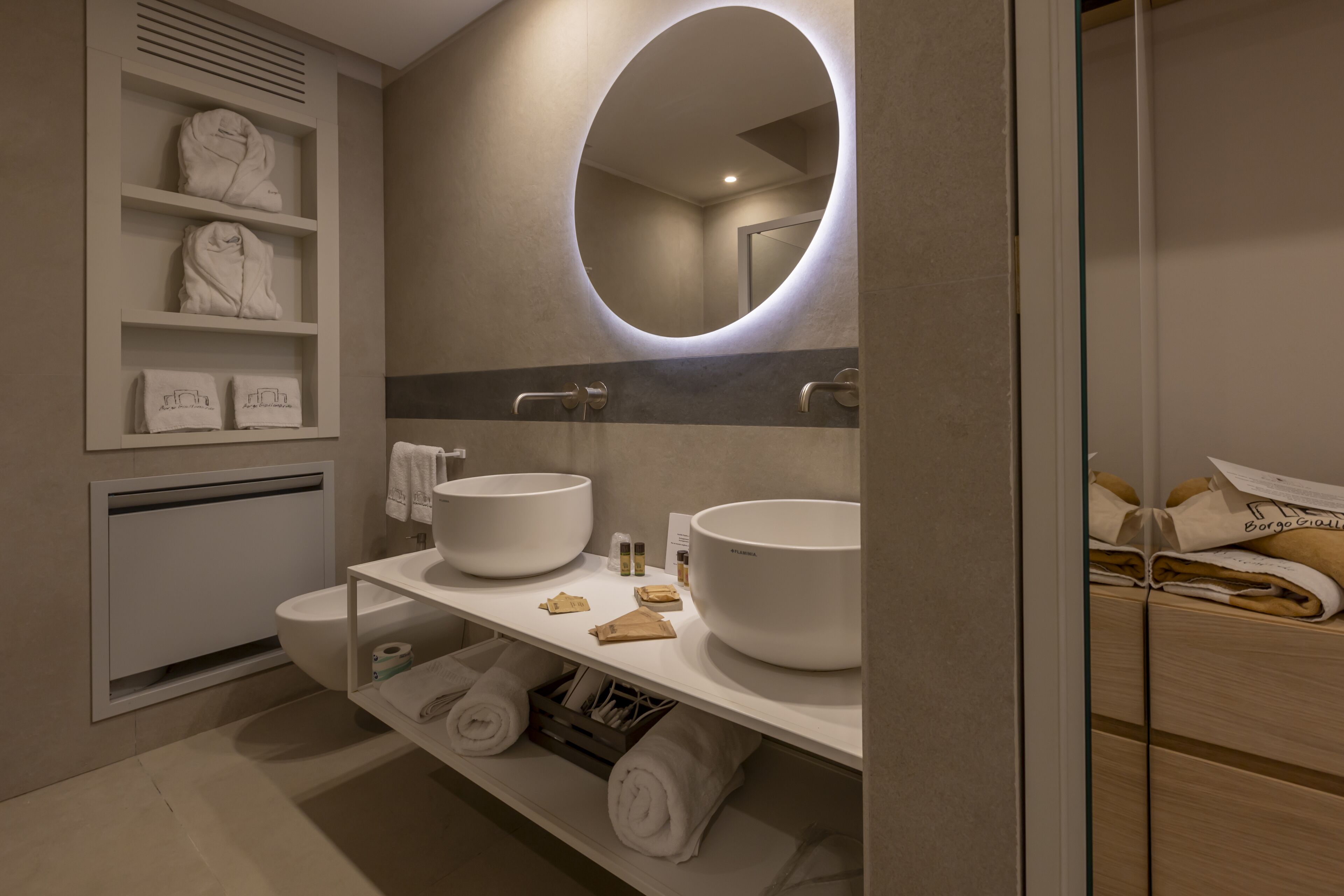 A modern bathroom with two large white sinks, a mirror with a circular light, shelves with towels, and a wooden cabinet.