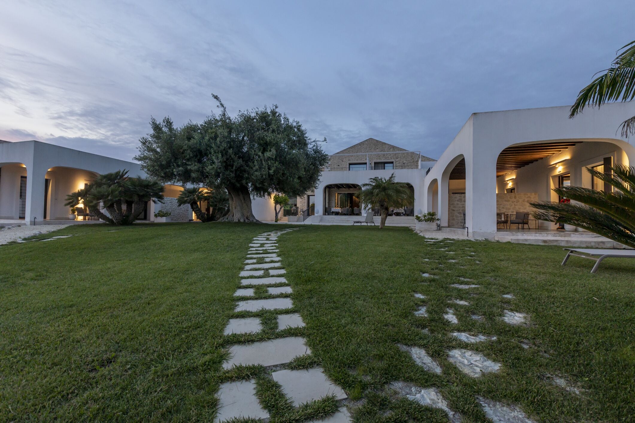 A modern house with a paved pathway leading to the entrance, surrounded by a well-maintained lawn and palm trees.