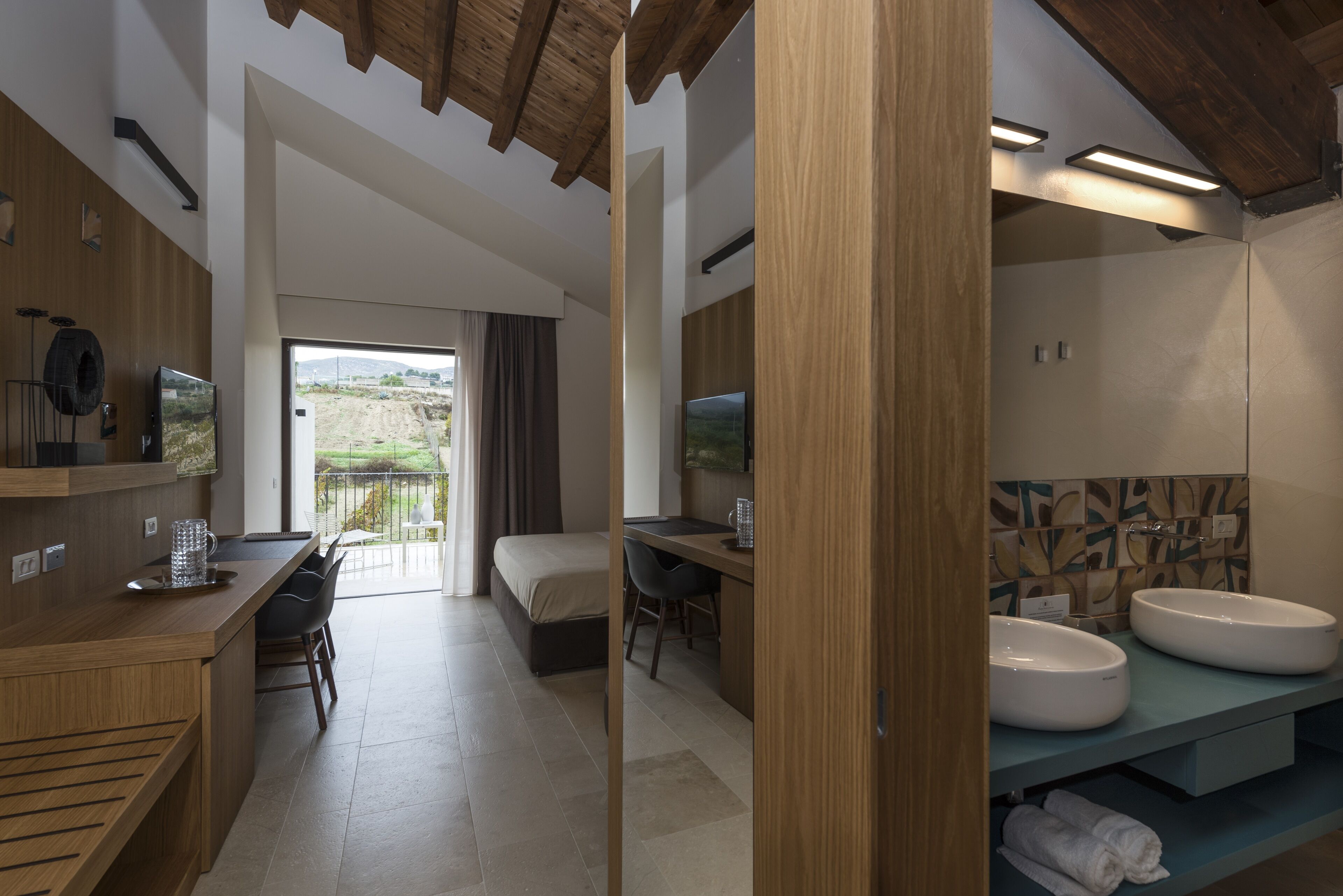A modern bathroom with two sinks, a shower, and a toilet. There is a wooden vanity with a mirror and a towel rack. The bathroom has a large window with a view of the outdoors.