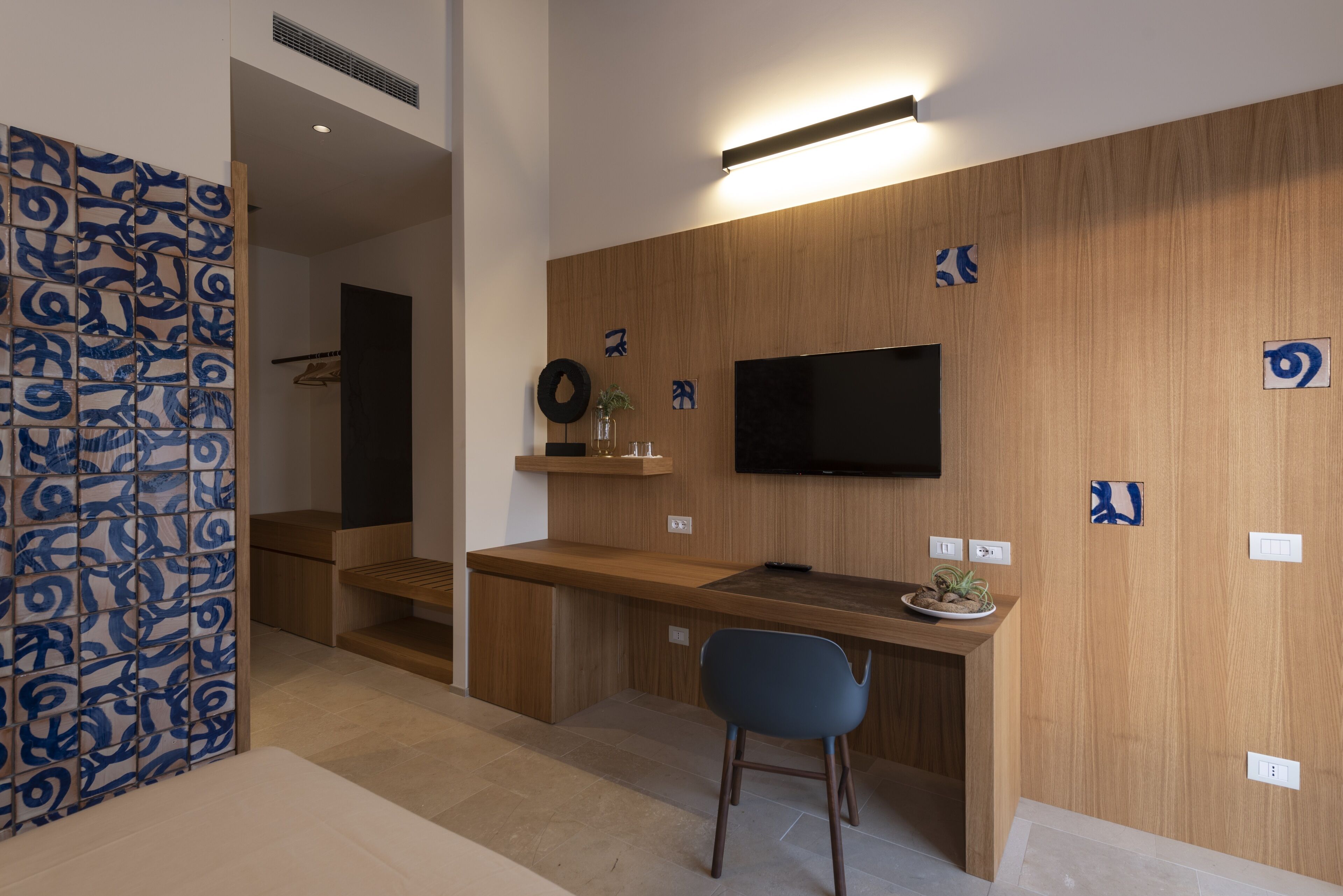Ein modernes Hotelzimmer mit Schreibtisch aus Holz, einem Flachbildfernseher an der Wand, einem blauen Sessel und einer dekorativen Wandverkleidung.
