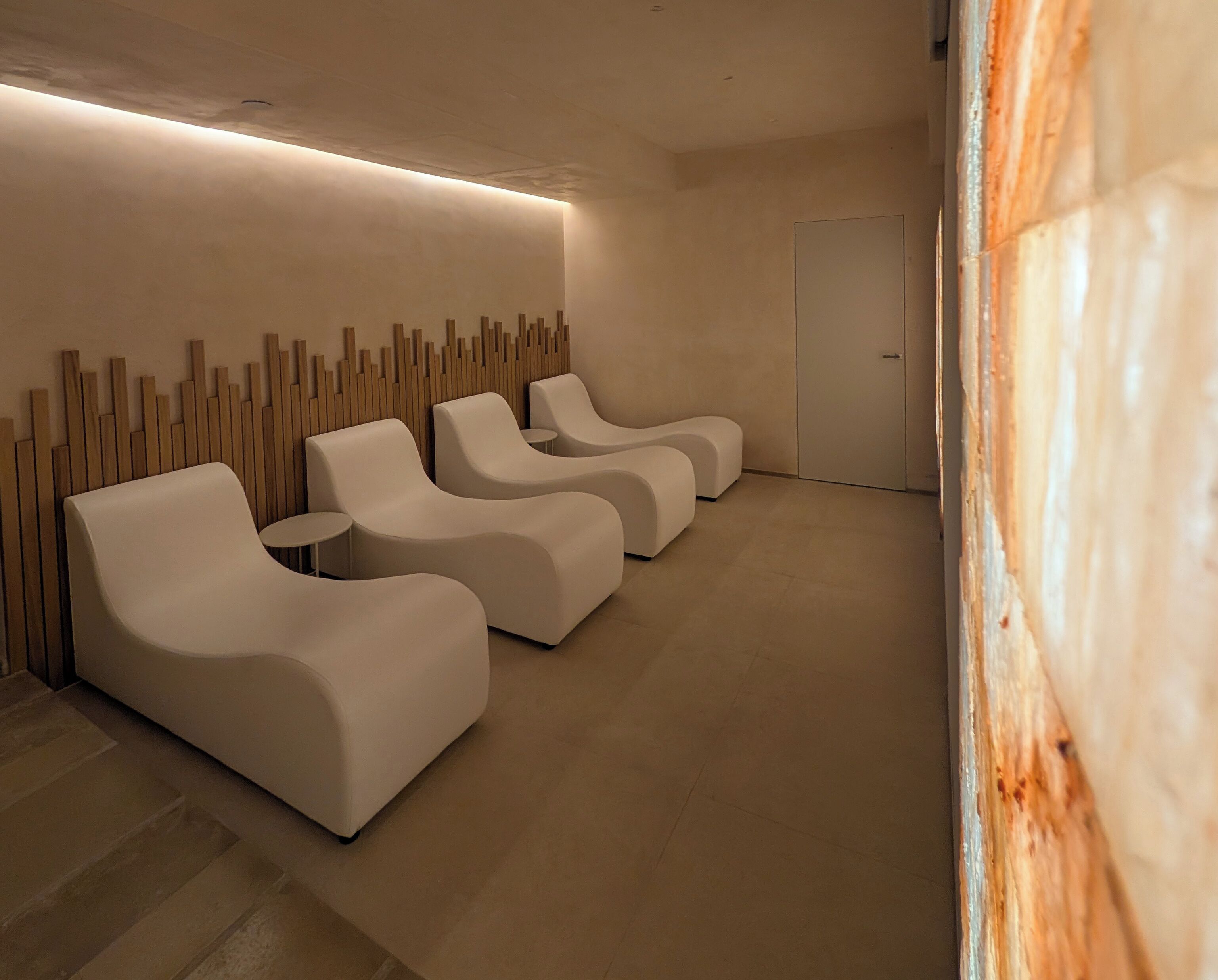 A modern spa room with three white lounge chairs, a wooden wall feature, and a door.
