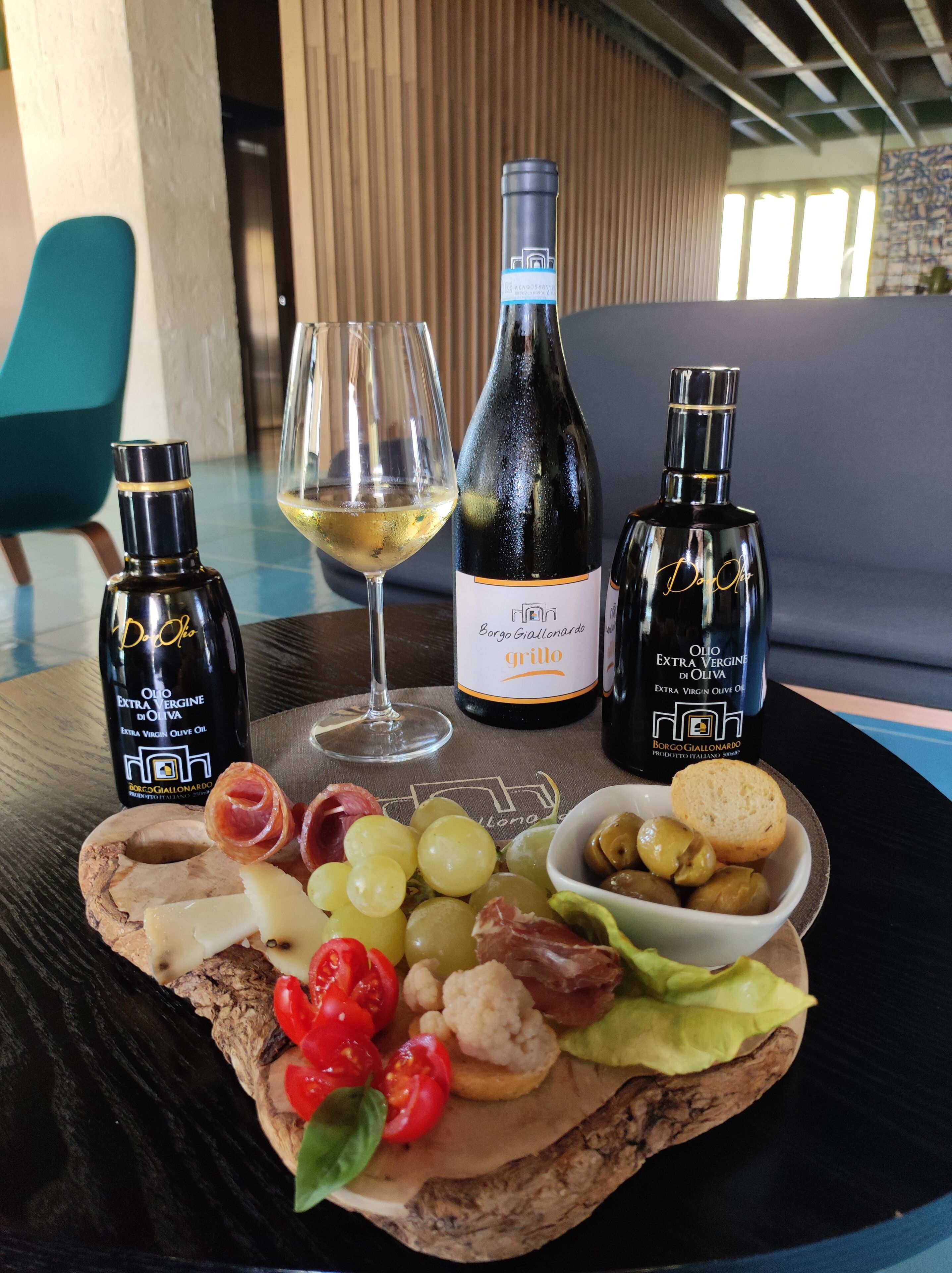 Ein Tisch mit einer Flasche Olivenöl, einer Weinflasche, einem Weinglas und einer Wurstplatte mit Käse, Trauben, Oliven und Brot.