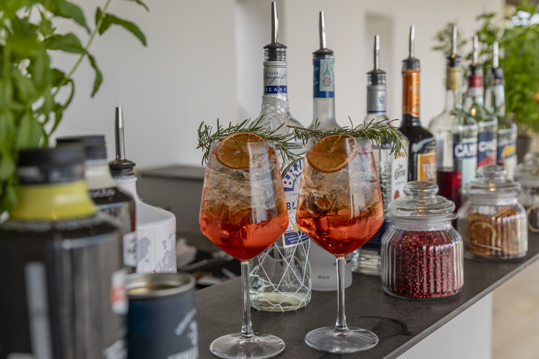 Zona de bar con dos vasos de cóctel rojo adornados con romero y rodajas de naranja, rodeada de botellas y frascos de condimentos.