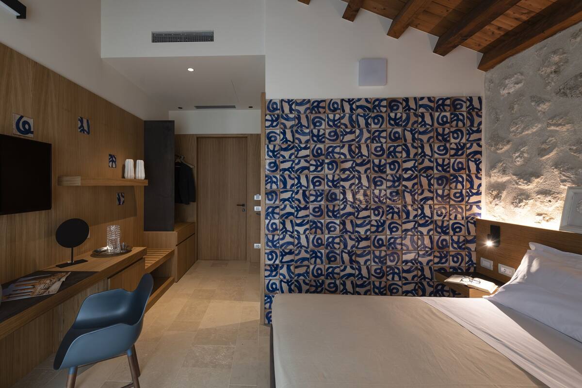 Une chambre d’hôtel moderne avec un bureau en bois, une chaise bleue, un lit avec du linge de maison blanc et un mur à motifs.