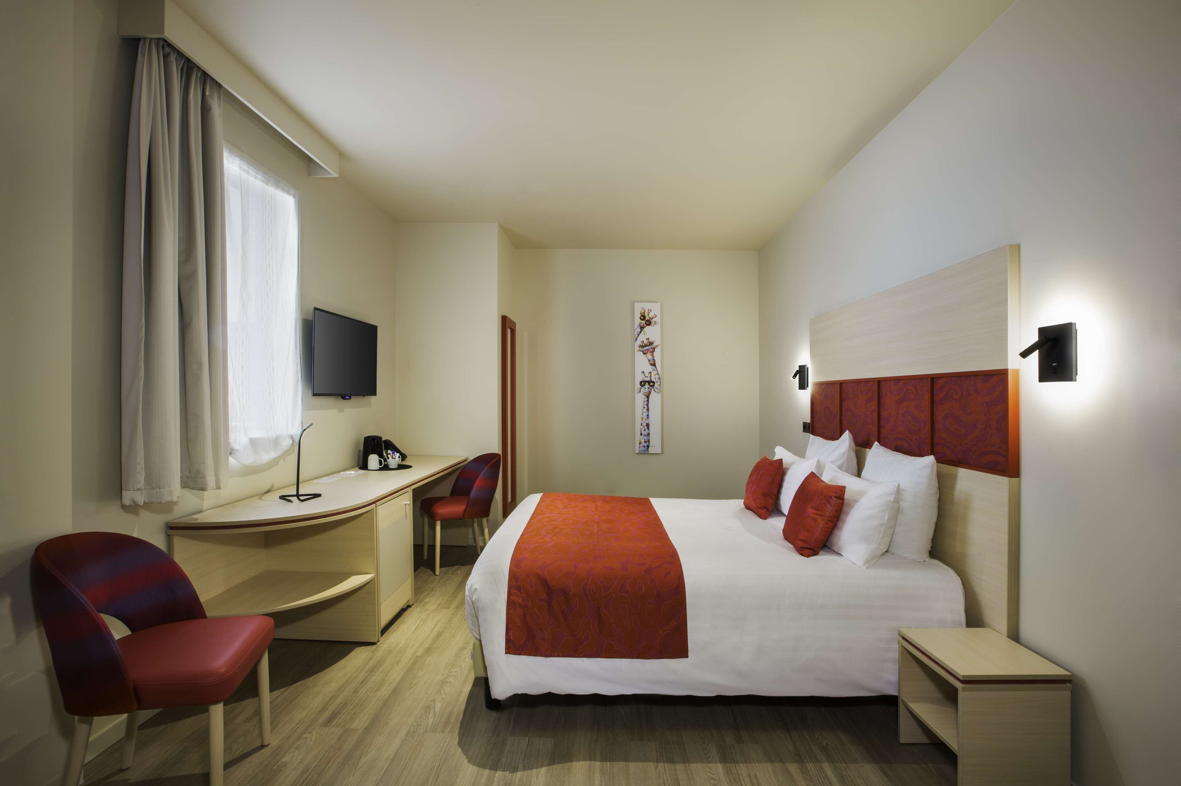 Habitación de hotel con cama, escritorio, silla, televisor y lámpara de pared.