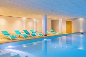 An indoor swimming pool with blue water, surrounded by white walls and ceiling, with several turquoise lounge chairs aligned along the pool's edge.