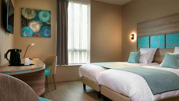 Une chambre d’hôtel comprenant un lit, un bureau, une chaise, une machine à café, un tableau, une fenêtre et un luminaire fixé au mur.