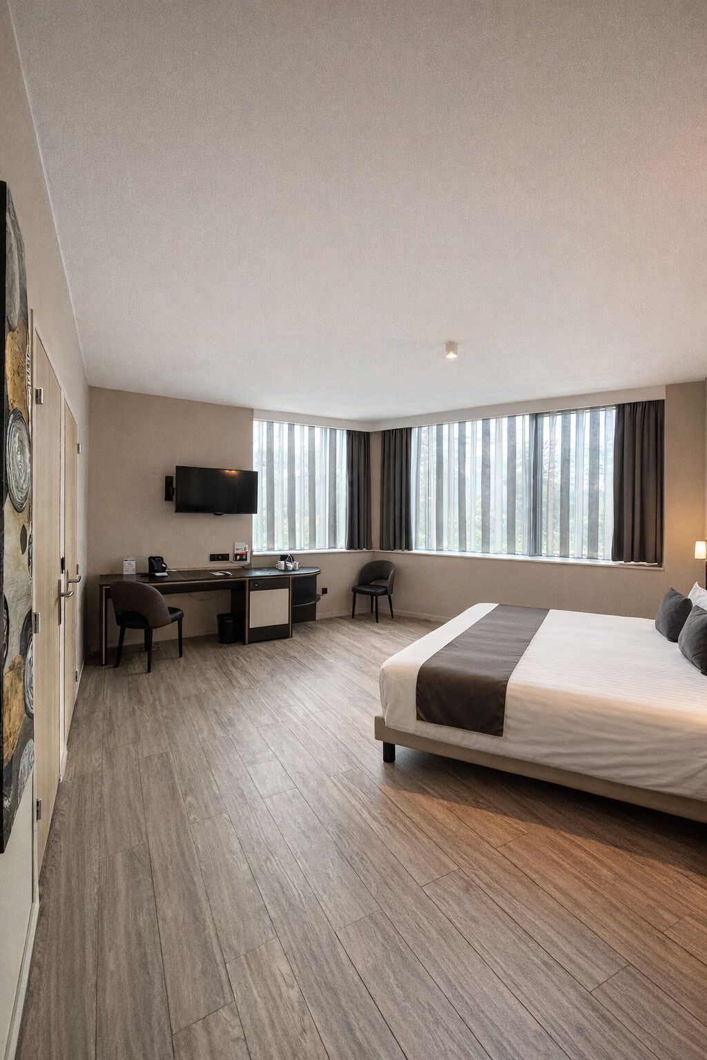 Habitación de hotel moderna con una cama grande, un escritorio con silla, un televisor de pantalla plana y un amplio ventanal con cortinas.