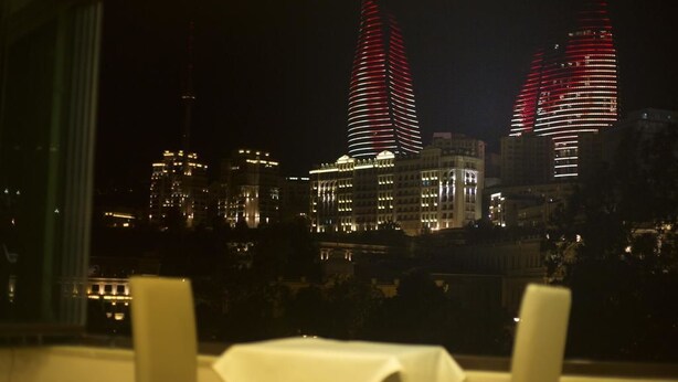 A nighttime cityscape with illuminated buildings, a table with a white tablecloth, and a window with curtains. - Town Gates Hotel (Baku)