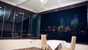 A dining area with a table and chairs, large windows, and a cityscape view at night. - Town Gates Hotel (Baku)